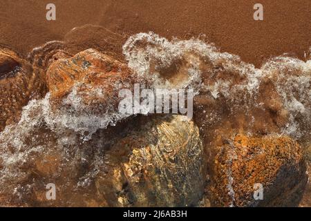 Direkt über dem Bild der kleinen Wellen, die in den Strand Rollen, übersät mit bunten Felsen und Felsbrocken Stockfoto