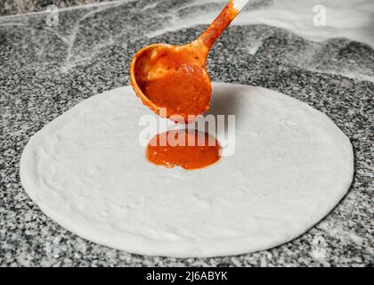 Der Koch gießt Tomatensauce über den gerollten Teig, Restaurantkonzept, den Prozess der Zubereitung italienischer Pizza Stockfoto