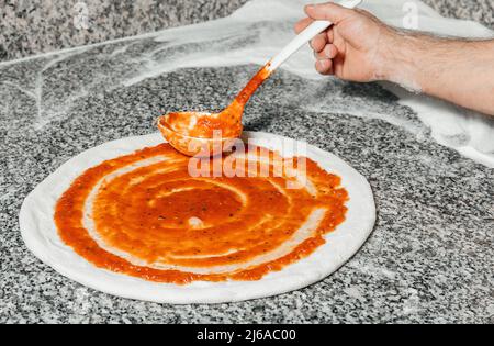 Der Koch gießt Tomatensauce über den gerollten Teig, Restaurantkonzept, den Prozess der Zubereitung italienischer Pizza Stockfoto