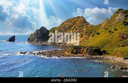 Wunderschöne malerische Küstenlandschaft mit rauer, rauer, grüner Felsküste, blauen Himmel und Sonnenstrahlen - Karitane, Neuseeland Stockfoto