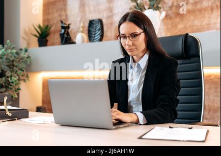 Intelligente kaukasische schöne Brünette Frau mit Brille, in einem formellen Anzug, Top-Manager, makler, Broker, sitzt an einem Schreibtisch im modernen Büro, arbeitet in einem Laptop, chatten online mit Kunden Stockfoto