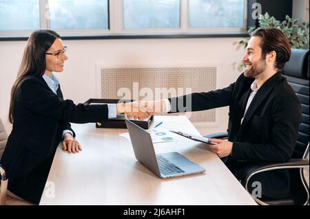 Selbstbewusste kaukasische Geschäftspartner geben sich bei Besprechungen oder nach erfolgreichen Verhandlungen die Hände, sitzen im modernen Büro. Einflussreiche Mitarbeiter unterzeichnen ein wichtiges Vertrags- oder Deal-, Collaboration-Konzept Stockfoto