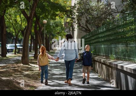 Rückansicht der glücklichen Großmutter, die Enkelkinder von der Schule nach Hause bringt und draußen auf der Straße läuft. Stockfoto