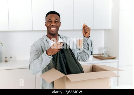 Lächelnder, attraktiver schwarzer Mann packte sein Paket aus und freute sich über eine lange erwartete Bestellung. Schöner Kerl Einkaufen in Internet-Shops, Kauf neuer Kleidung online, Online-Shopping, Lieferkonzept Stockfoto