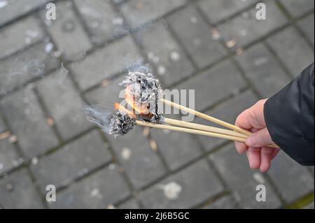 Die Hand einer erwachsenen Frau hält drei brennende Papierblumen vor einem Winterpark. Das schwarz verkohlte Papier raucht Stockfoto