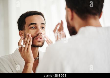 Junger Mann, der transparente Flecken unter seinen Augen auflegte, während er im Bad gegenüber dem Spiegel stand Stockfoto