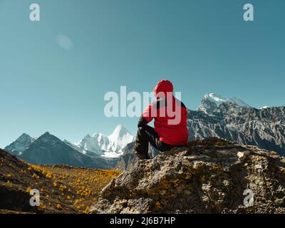 Rückansicht eines asiatischen Mannes, der auf einem Felsen mit dem Yangmaiyong (oder auf Tibetisch Jampayang)-Berggipfel in der Ferne in Yading, Landkreis Daocheng, sic sitzt Stockfoto