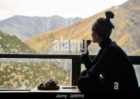 asiatische Frau, die auf dem Balkon sitzt und Tee trinkt und die Berglandschaft mit herbstlicher Vegetation genießt Stockfoto