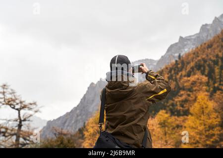 Rückansicht eines asiatischen männlichen Touristen, der mit dem Handy ein Landschaftsfoto von Berg- und Herbstlaub gemacht hat Stockfoto