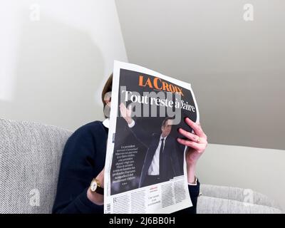 Paris, Frankreich - 25. Apr 2022: Frau liest auf einer gemütlichen Couch die neueste Zeitung La Croix, die mit Frankreichs amtiertem Präsidenten Emmanuel Macron berichtet, nachdem sie Marine Le Pen für eine zweite fünfjährige Amtszeit geschlagen hatte Stockfoto