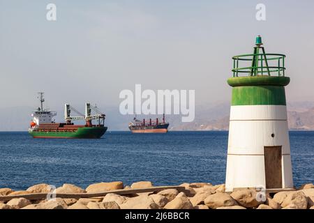 Kleiner Leuchtturm an der Küste von Aqaba, Jordanien, mit Frachtschiffen im Hintergrund. Dieser geschäftige Hafen liegt am Roten Meer und ist der einzige Seehafen in Jo Stockfoto