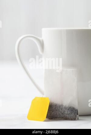 Teebeutel mit gelbem, unbeschrifttem Anhänger von schwarzem Tee neben Porzellanbecher auf weißem Hintergrund. Stockfoto