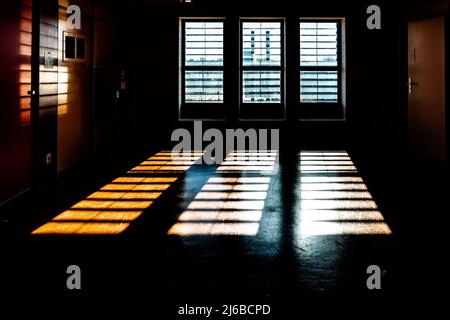 Helles Licht dringt durch die Fenster ein und wirft dunkle Schatten auf den Boden. Bars sind vor den Fenstern zu sehen, wie in einem Gefängnis. Das Bild ev Stockfoto