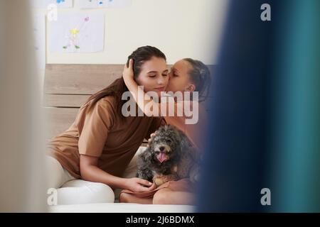 Mädchen küsst Mutter auf der Wange, wenn sie auf dem Bett mit einem kleinen Hund sitzen Stockfoto