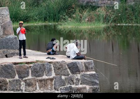 Vellore, Tamil Nadu, Indien - September 2018: Menschen, die am Rande einer Steinmauer des Vellore Fort sitzen und mit Holzstäben fischen. Stockfoto