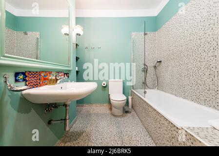 Badezimmer mit hängendem Waschbecken aus weißem Porzellan, großem Spiegel mit grünem Rahmen und verglaster Badewanne mit Kachelfliesen Stockfoto