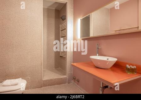 Badezimmer mit weißem Porzellanwaschbecken auf orangefarbener Glasplatte, Spiegel auf der Arbeitsplatte, gefliesten Wänden und begehbarer Dusche Stockfoto