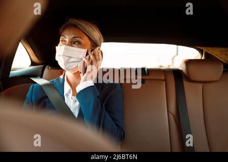 Reife asiatische Geschäftsfrau trägt Gesichtsmaske reden auf Handy im Auto Stockfoto