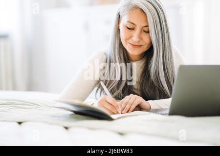 Reife asiatische Frau Notizen beim Arbeiten mit Laptop zu Hause Stockfoto