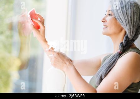 Reife asiatische Frau mit grauen Haaren Reinigung Fenster mit Spray zu Hause Stockfoto