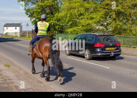 Auto überholen Frau Reiten Pferd auf Landstraße trägt hohe Sichtbarkeit gelben Tabard mit "Bitte Pass breit und langsam prangt. Stockfoto