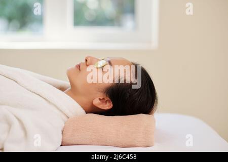 Ruhige, entspannte junge Frau, die im Spa-Salon mit Gurkenscheiben auf geschlossenen Augen auf dem Bett liegt Stockfoto
