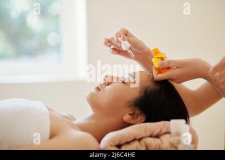 Kosmetikerin, die einige Tropfen feuchtigkeitsspendenden Öls auf das Gesicht einer jungen Frau setzt Stockfoto