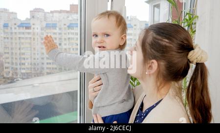 Junge Mutter hält ihren kleinen Sohn und versucht, das Fenster in einem der oberen Stockwerke zu öffnen. Baby in Gefahr. Kindersicherheit Stockfoto