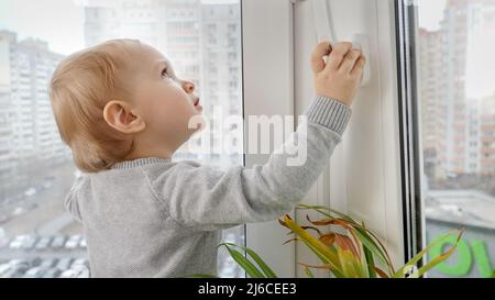 Kleines Baby, das den Knopf drückt und den Fenstergriff zieht, um das Fenster zu öffnen. Kind in Gefahr zu Hause. Stockfoto