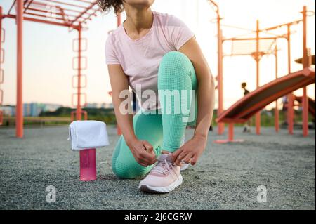 Beschnittenes Bild einer Sportlerin, die Schnürsenkel vor einem schweren Training auf Fitnessgeräten auf dem Sportplatz eines Stadtparks auf einem warmen Su bindet Stockfoto