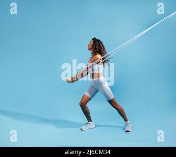 Weibchen beim Ausarbeiten mit Gummiband auf blauem Hintergrund. Schlanke, athletische Frau, die mit einem Widerstandsband an der Kernstärke arbeitet. Stockfoto