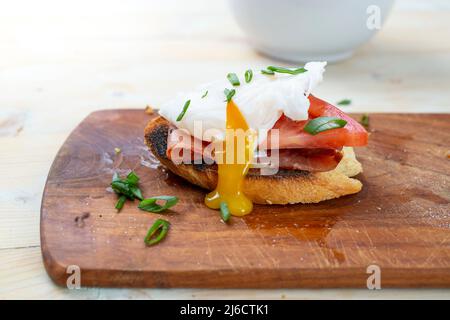 Pochiertes Ei mit tropfendem Eigelb, Schinken- und Tomatenscheibe, auf Toastbrot, Seitenansicht aus nächster Nähe Stockfoto