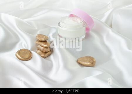 Glasglas mit sichtbarer weißer Creme und geöffnetem rosafarbenen Deckel neben einem Stapel goldener Steine Stockfoto