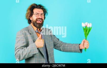 Glücklicher bärtiger Mann mit Blumen, die den Daumen nach oben zeigen. Geschäftsmann mit Tulpenstrauß zum Geburtstag. Stockfoto