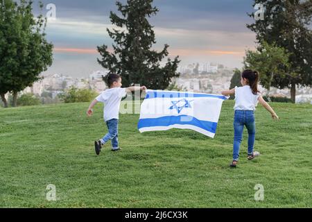 Kleine Kinder laufen mit israelischer Flagge. Rückansicht kleines Mädchen und Junge, die bei Sonnenuntergang mit der israelischen Flagge einen Hügel hochlaufen Stockfoto