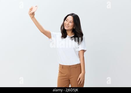 Lasst mich ein Selfie machen. Attraktive fröhliche asiatische junge Frau in weißem Grund T-Shirt tun Schuss mit Telefon posiert isoliert auf weißem Studio Hintergrund Stockfoto