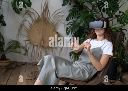 Aufgeregte junge Frau, die sich auf einen Stuhl lehnt und ein vr-Headset trägt, spielt in virtuellen Spielen im Heimgarten Stockfoto