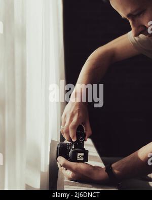 Der junge Mann fotografiert an einem sonnigen Tag mit einer Kamera in der Nähe des Fensters. Konzept der Seitenansicht. Stockfoto