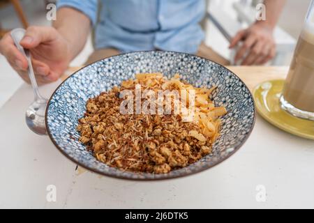 Schüssel mit Granulat und Kokosnuss- und Schokoladenchips für ein gesundes Frühstück. Männliche Hand mit einem Löffel und einem Teller mit Müsli in einem Café am Morgen, c Stockfoto