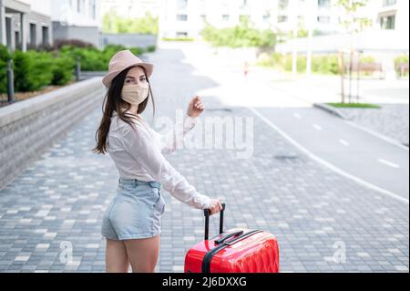 Eine schöne junge kaukasische Frau in einer medizinischen Maske reist mit einem roten Koffer durch eine leere Stadt. Das Mädchen geht mit einem großen Gepäck in den Urlaub Stockfoto