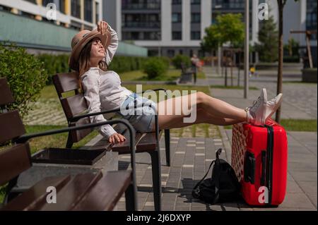 Eine schöne junge Frau sitzt auf einer Bank und legt ihre Beine auf einen roten Koffer vor der Tür Stockfoto