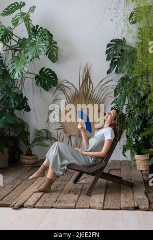 Junge, entspannte, verträumte Frau, die einen Papierventilator verwendet, während sie sich im schönen, erfrischenden Hausgarten entspannt Stockfoto