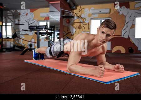 Selbstbewusster, muskulös junger Mann, der Sportkleidung trägt und beim Training auf dem Boden im Fitnessstudio eine Plankenposition einnimmt Stockfoto