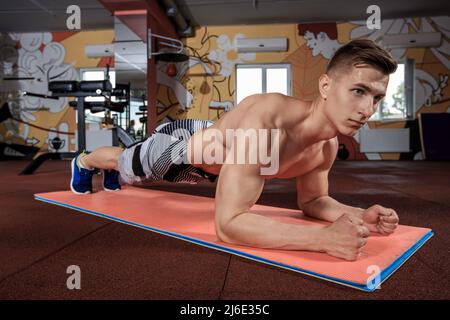 Selbstbewusster, muskulös junger Mann, der Sportkleidung trägt und beim Training auf dem Boden im Fitnessstudio eine Plankenposition einnimmt Stockfoto