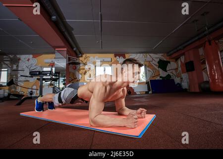 Selbstbewusster, muskulös junger Mann, der Sportkleidung trägt und beim Training auf dem Boden im Fitnessstudio eine Plankenposition einnimmt Stockfoto