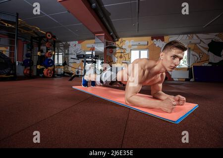 Selbstbewusster, muskulös junger Mann, der Sportkleidung trägt und beim Training auf dem Boden im Fitnessstudio eine Plankenposition einnimmt Stockfoto