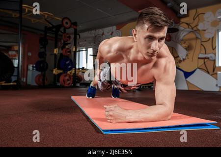 Selbstbewusster, muskulös junger Mann, der Sportkleidung trägt und beim Training auf dem Boden im Fitnessstudio eine Plankenposition einnimmt Stockfoto