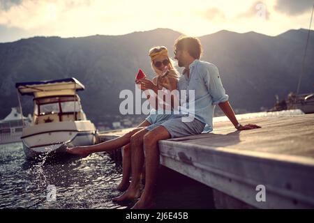 Pärchen, die sich klein unterhalten und Wassermelone am Wasser genießen, auf einem hölzernen Steg sitzen. Frau spritzt Wasser mit ihrem Bein. Liebe, Urlaub, Lifestyle conce Stockfoto