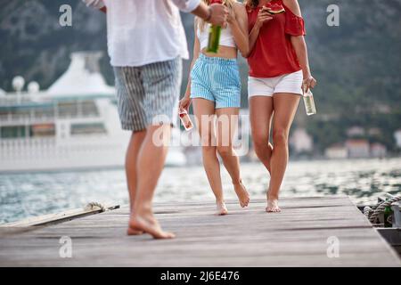 Eine Gruppe von Freunden auf einem hölzernen Steg, die mit einem Glas Bier und einem Stück Wassermelone unterwegs sind. Spaß, Zweisamkeit, Urlaubskonzept. Stockfoto