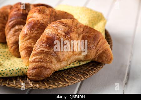 Frisches Croissant, auf gelber und grüner Serviette und Weidenkorb, auf rustikalem Hintergrund aus weißem Holz. Low-Angle-Ansicht mit Platz für Text. Stockfoto